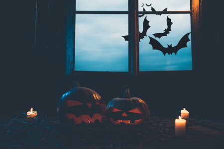Halloween - pumpkins and candles in an abandoned wooden house on leaves and wooden boards with a warm and cold glow, against the background of a window with a mystic sky and batsの写真素材