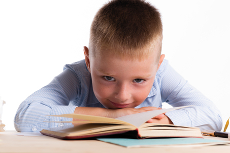 Cute little school boy with huge smile sitting at his desk on white background. Happy intelligent children in shirt with blue eyes, lies on table with bookの写真素材