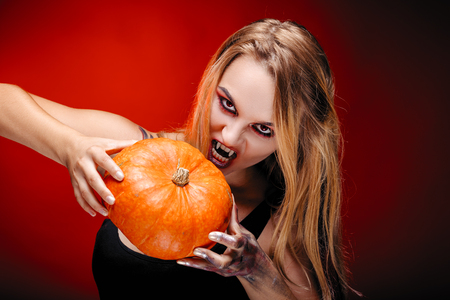 Halloween girl vampire with red eyes red lips and dark red background. Scary woman trick or treat time, halloween concept. Female makeup for holiday with pumpkin in handsの写真素材