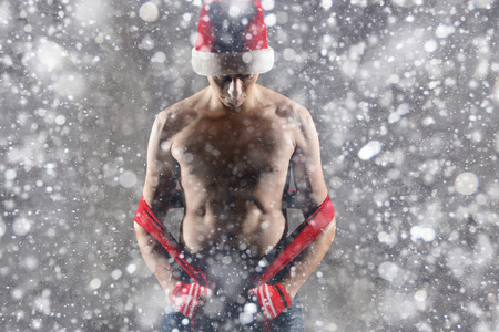 Christmas gift concept. Man in santa hat bring gift for you. Attractive man hold box.  Christmas and new year holiday celebration. Happy man under snowflakes with nice torso wear hat, red scarfの写真素材
