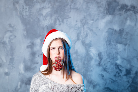 Merry Christmas and happy new year! Adorable girl with ball toy in mouth looks very puzzled. Close portrait on gray background. Girl in santa hat and sweaterの写真素材