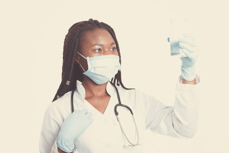 Female american african doctor, nurse woman wearing medical coat with stethoscope, mask and gloves. Doctor holding glass of water. Serious excited for success medical worker posing on light backgroundの写真素材