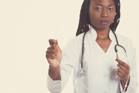 Female american african doctor, nurse woman wearing medical coat with stethoscope and needle dropper. Serious excited for success medical worker posing on light backgroundの写真素材