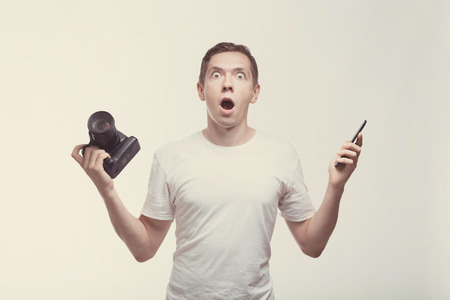 Emotional Man with camera isolated on light background. Young man holding camera and smartphone....