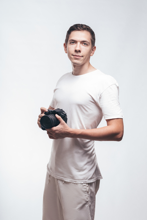 Serious Man with camera isolated on light background. Young man holding digital camera and look on you. Lifestyle, travel and technology concept. Happy boy in white t-shirt love work and dslr cameraの写真素材