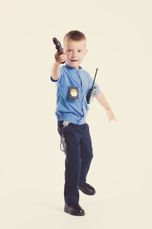 Cute little police boy with smile on face and gun on white background. Intelligent cool children in police suit with blue eyes and weaponの写真素材