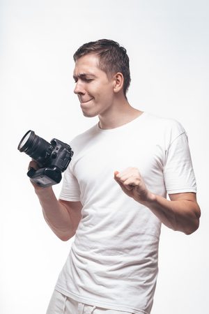 Emotional Man with camera isolated on light background. Young man holding camera with victory dancing. Lifestyle, travel and technology concept. Glad boy in white t-shirtの写真素材