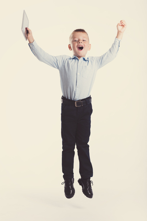 Happy little boy in suit jump and hold tablet gadget. Children portrait. Back to school. Stylish man in fashionable suit shirt and pants with blue eyesの写真素材