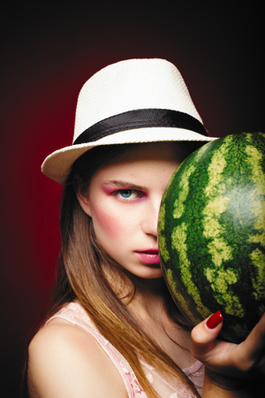 Pretty serious girl with pink make up, wearing jeans, hat and top, posing at red studio background, holding watermelon, emotionally standの写真素材
