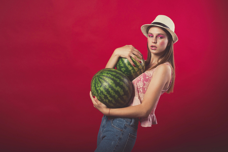 Confident Attractive girl with pink make up, wearing jeans, hat and top, posing at red studio background, holding few watermelon, emotionally standの写真素材