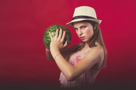 Confident Attractive girl with pink make up, wearing jeans, hat and top, posing at red studio background, holding watermelon, emotionally standの写真素材