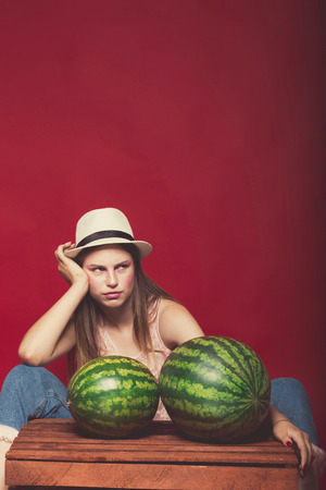 Gorgeous girl with pink make up, wearing jeans, hat and top, posing at red studio background near wooden box, holding watermelon, emotionally sittingの写真素材
