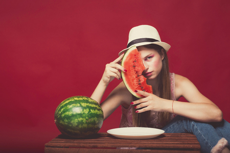 Lovely girl with pink make up, wearing jeans, hat and top, posing at red studio background near wooden box, holding slice watermelon, emotionally sittingの写真素材