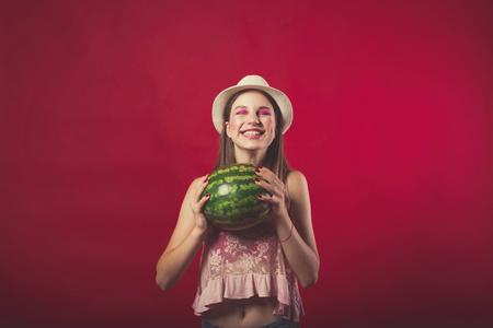 Gorgeous girl with pink make up, wearing jeans and top, posing at red studio background, holding watermelon, looking at camera and smileの写真素材