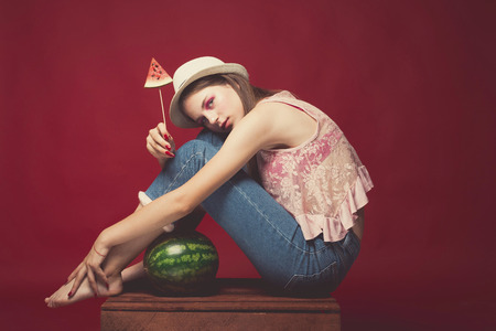 Attractive Lovely girl with pink make up, wearing jeans, hat and top, posing at red studio background sitting on wooden box, holding slice watermelon, emotionally lookの写真素材