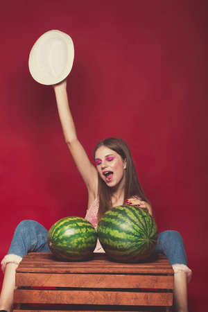 Lovely girl with pink make up, wearing jeans, hat and top, posing at red studio background near wooden box, holding few watermelon, looking at camera and smile emotionallyの写真素材