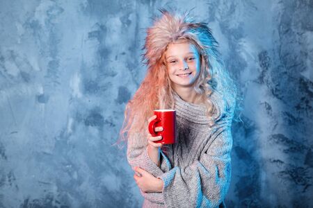 Merry Christmas and happy new year! Adorable young female hold red cup. Close portrait on gray background. Girl in fur hat and sweater dreaming looks pleasedの写真素材
