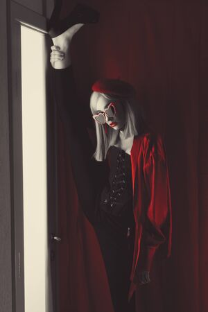 French girl in red beret with red glasses with heart and big beautiful eyes in an interior loft studio lifts her leg up in standing twine. Woman in red jacket. Fashionable female with white hairの写真素材