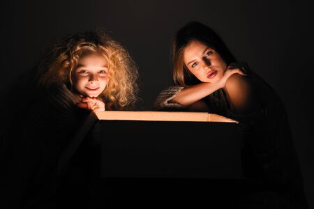 Merry Christmas and happy new year! happy girl near big present open it, and light from it blessing on her. Miracle in dark holiday time. Sisters looking in box present from santa claus or parents.の写真素材