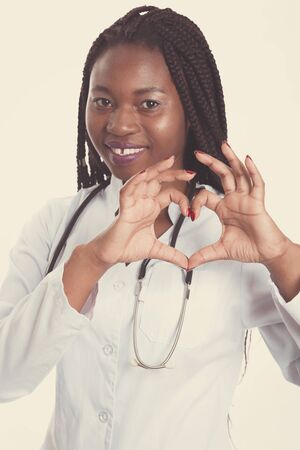 Female american african doctor, nurse woman wearing medical coat with stethoscope doing heart with hands. Happy excited for success medical worker posing on light background isolatedの写真素材
