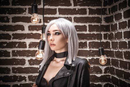 Girl in a leather jacket with white hair in an interior loft studio among light bulbs. The girl in the bra. Fashionable Female stands at the brick. Fashionable female with gray hairの写真素材