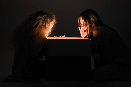 Merry Christmas and happy new year! happy girl near big present open it, and light from it blessing on her. Miracle in dark holiday time. Sisters looking in box present from santa claus or parents.の写真素材