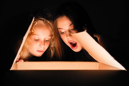 Merry Christmas and happy new year! happy girl near big present open it, and light from it blessing on her. Miracle in dark holiday time. Sisters looking in box present from santa claus or parents.の写真素材
