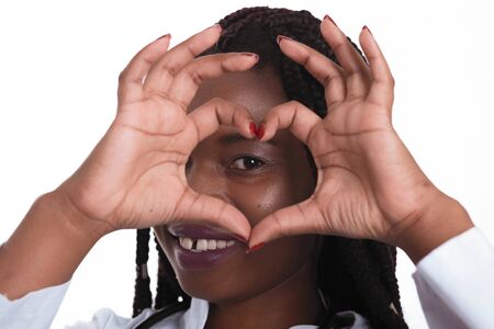 Female american african doctor, nurse woman wearing medical coat with stethoscope doing heart with hands and looking through it. Happy excited for success medical worker posing on light background isolatedの写真素材
