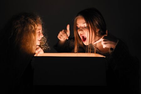Merry Christmas and happy new year! happy girl near big present open it, and light from it blessing on her. Miracle in dark holiday time. Sisters looking in box present from santa claus or parents.の写真素材