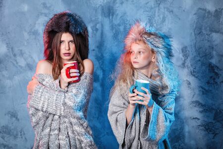 Merry Christmas and happy new year! Adorable sisters holds red and blue cups. Close portrait on gray background. Girls in fur hat and sweater looks coldの写真素材