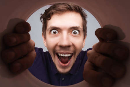 Man looking inside cup or rounded tube with hands on it. Close-up shot of happy shocked young man with round eyes and open mouth smile.の写真素材