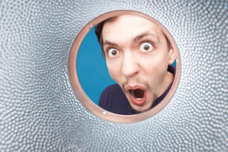 Man looking inside cup or rounded metal tube. Close-up shot of shocked young man with round eyes and open mouth.の写真素材