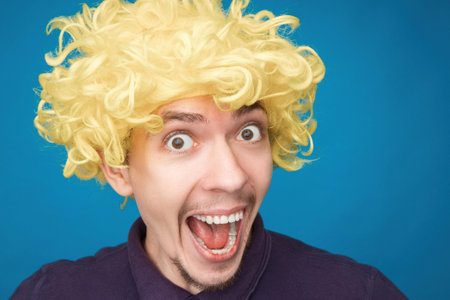 Funny portrait of a man with happy emotion on his face in the studio on blue background. Man wearing wig. Yellow hairの写真素材