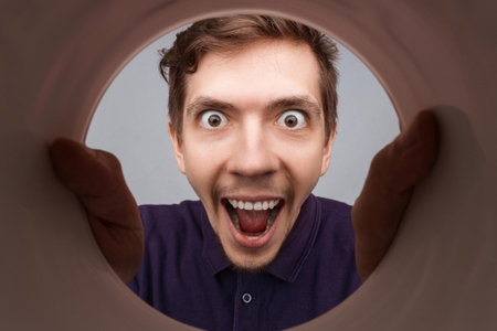 Man looking inside cup or rounded tube with hands on it. Close-up shot of happy shocked young man with round eyes and open mouth smile.の写真素材