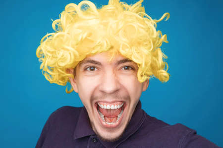 Funny portrait of a man with happy emotion on his face in the studio on blue background. Man wearing wig. Yellow hairの写真素材