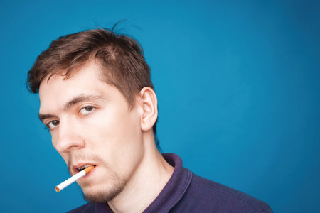 Handsome young man smoking cigarette. one caucasian stylish man smoking cigarettes portrait on blue backgroundの写真素材