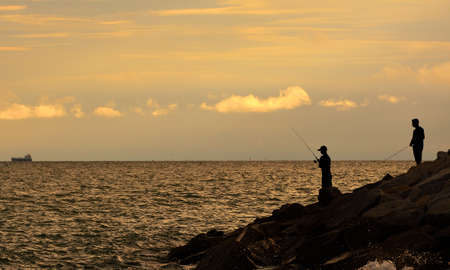 Black silhouette of two men holding a fishing rod by the sea at  sunsetの写真素材