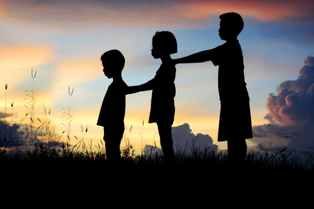 Black silhouette of three children standing shoulder to shoulder. There is a sky at sunset.の写真素材