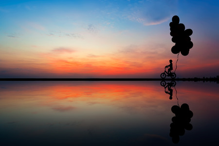 Silhouette of children on bicycle with air balloons at sunset skyの写真素材