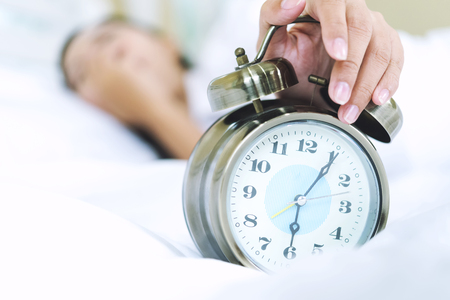 Sleepy young woman in bed with eyes closed extending hand to alarm clock. Lazy day concept.の写真素材