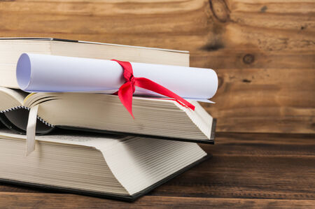 A parchment diploma scroll, rolled up with red ribbon on stack of book on wood backgroundの写真素材