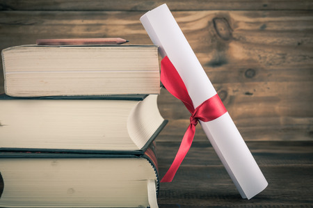 A parchment diploma scroll, rolled up with red ribbon beside a stack of books on wood background with vintage filterの写真素材