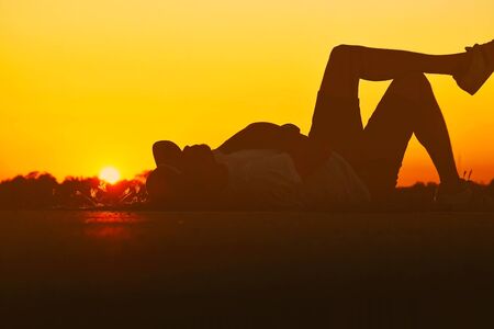Silhouette of young asian people lying on the concrete floor for recreation near to natural water pond at sunset time isolated with orange color of sky light.の写真素材
