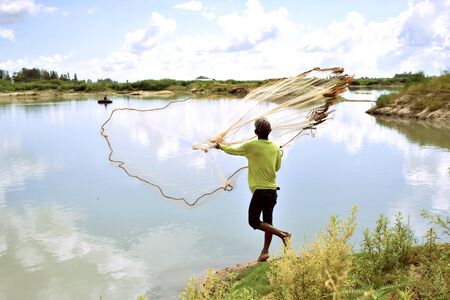 Back view of fisherman while throwing fishing net in nature water pond. Rural life of people about...