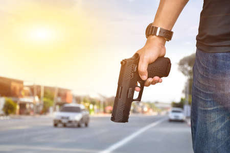 Man wears jeans holds an automatic 9mm pistol, cropped shot of man holding shotgun in hand with blurred street bokeh background. Concept for robbery on the road and security profession in the city.の写真素材