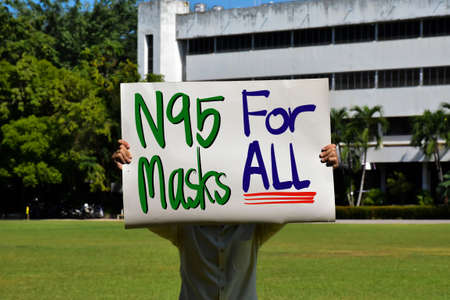 Asian man holds protesting white paper which has texts 'N95 Masks For All' in the middle green grass...