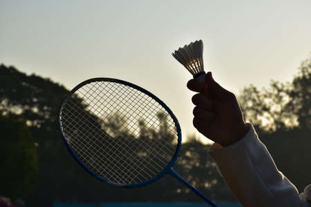 Badminton racket and new badminton shuttlecock holding in hand of player, blurred outdoor and sunset...