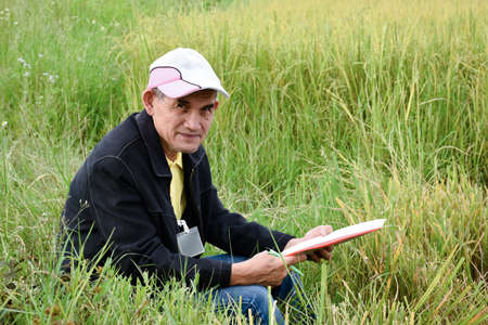 Portrait of Asian senior volunteer who holds white paper and pen in hand, sits on grass near rice...