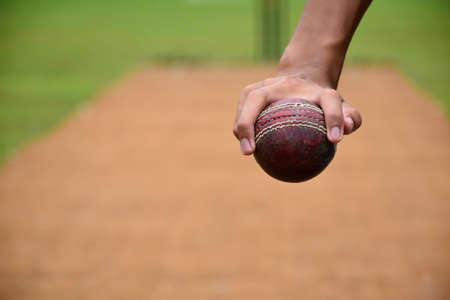Right hand of cricketer holds old red leather cricket ball, soft and selective focus on hand, blurred cricket court background.の写真素材