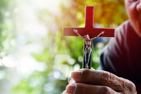 Closeup wooden cross which has a metal statue of crucified Jesus is in the hands of an asian eldery Catholic while praying in a local church, soft and selective focus.の写真素材